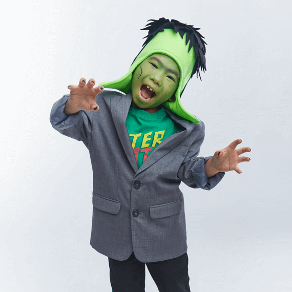 A child with green face paint with a frankenstein hat, a green shirt and a dark grey suit jacket on