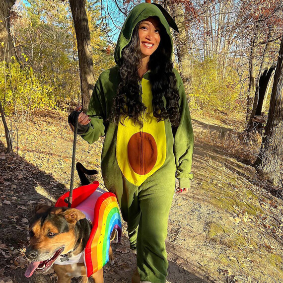 A female dressed in an avocado onesie Halloween costume. Her dog is dressed in a rainbow costume. 