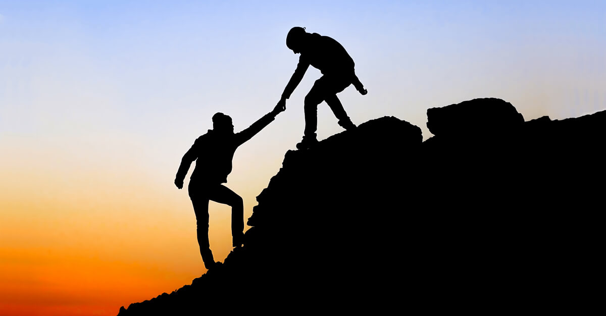 Photo of the sillouttes of two individuals climbing with one person pulling the other up.