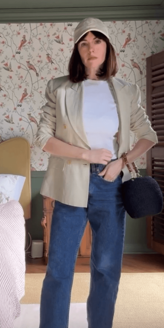 A female wearing a baseball cap, blazer, top, jeans and a black handbag.