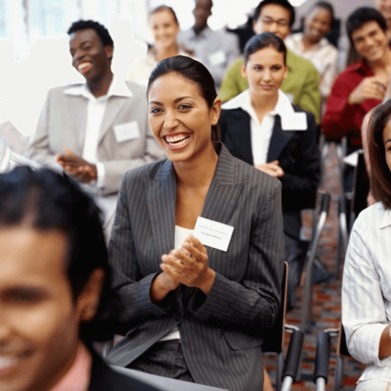 A group of corporate professionals at an event. A female is wearing a pin-stripped jacket with a name tag and is clapping.