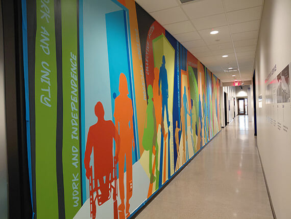 Photograph of the mural in the hallway from the left at GESMN's career campus. The mural is very colorful and shows people walking through doorways.