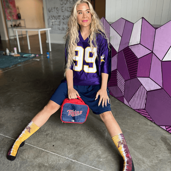 A female with a Vikings purple football jersey on, who is holding a Twins red, white and blue lunch box.