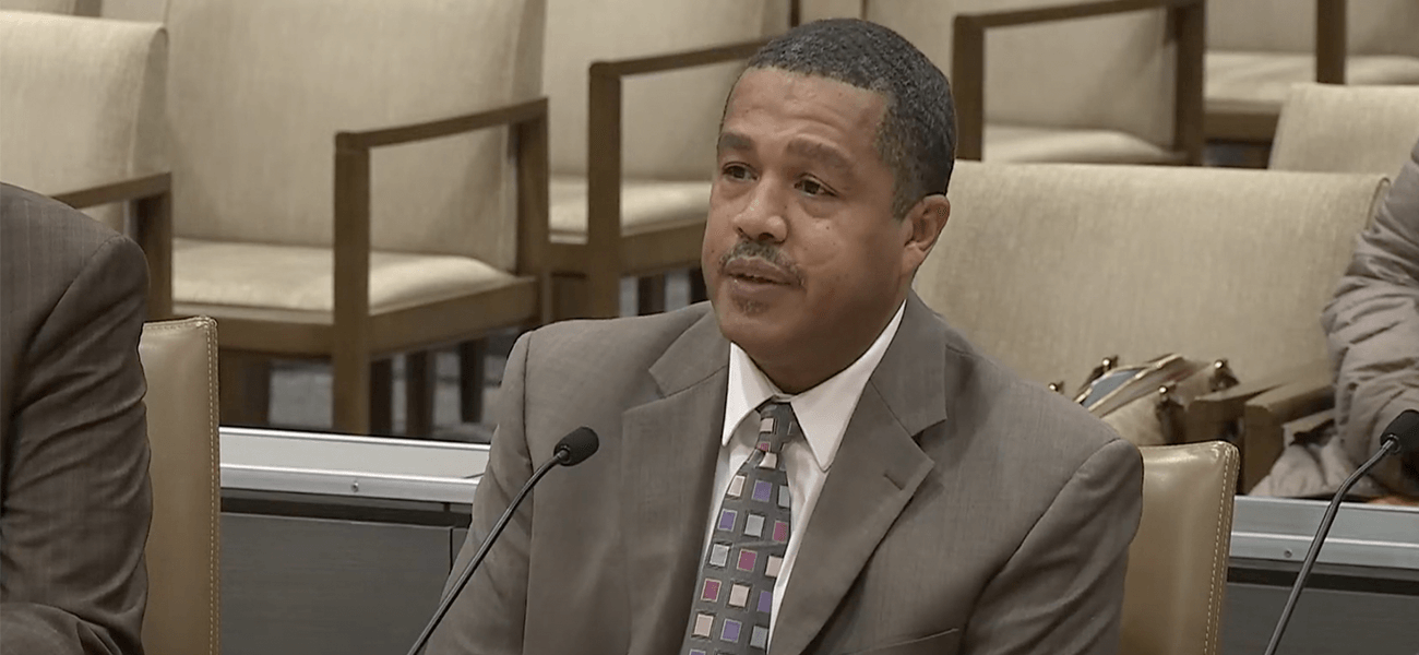 Individual testifying at the Capitol, wearing a suit and tie, with a microphone in front.