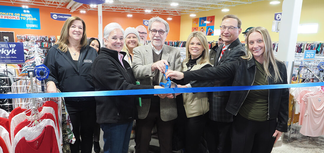 A group of people stands together indoors at a ribbon-cutting event. They are holding a large blue ribbon, with scissors poised to cut. 