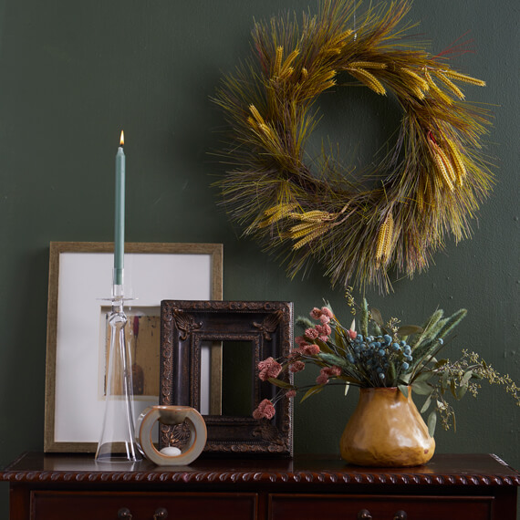 Two picture frames, a candle and a vase with flowers on a table. A wreath is hanging on a green wall.