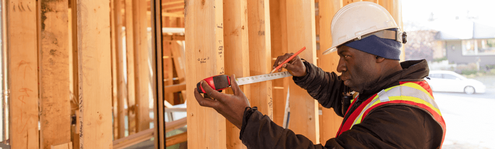 A male in construction gear, holding up a tape measure on wood siding